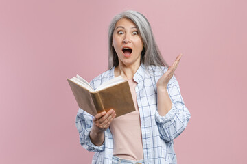 Shocked gray-haired asian woman wearing basic white checkered shirt standing holding in hands reading book spreding hands looking camera isolated on pastel pink colour background, studio portrait.