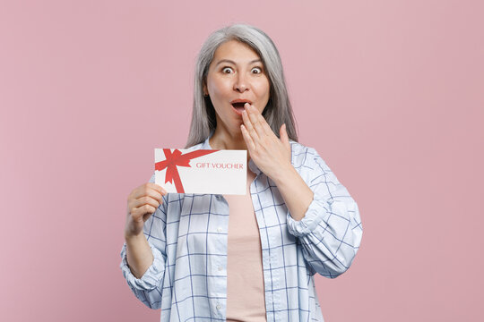 Shocked gray-haired asian woman wearing basic white checkered shirt hold in hand gift certificate covering mouth with hand looking camera isolated on pastel pink colour background, studio portrait.
