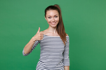 Smiling pretty funny attractive young brunette woman 20s wearing striped casual clothes posing standing showing thumb up looking camera isolated on green color wall background, studio portrait.