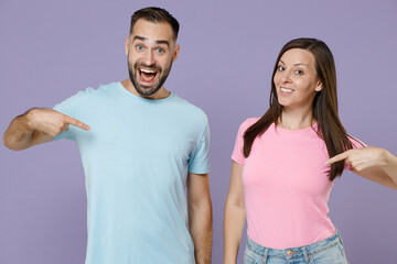 Smiling excited young couple two friends man woman in blue pink empty blank design t-shirts standing pointing index fingers on themselves isolated on pastel violet color background studio portrait.