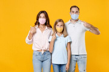 Young parents mom dad with child kid daughter teen girl in basic t-shirts pointing index fingers on face mask to safe from coronavirus virus covid-19 isolated on yellow background. Family day concept.