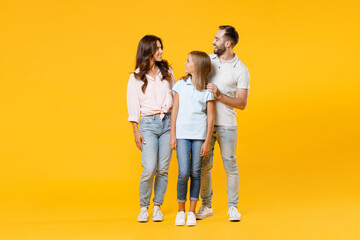 Full length portrait cheerful young parents mom dad with child kid daughter teen girl in t-shirts hugging looking at each other isolated on yellow background. Family day parenthood childhood concept.