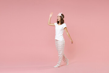 Fototapeta na wymiar Full length side view portrait funny young woman in white pajamas home wear sleep mask waving greeting with hand as notices someone rest at home isolated on pink background. Relax good mood concept.