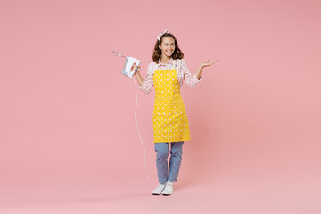 Full length portrait of smiling young woman housewife 20s wearing yellow apron hold mixer spreading hands while doing housework isolated on pastel pink colour background studio. Housekeeping concept.