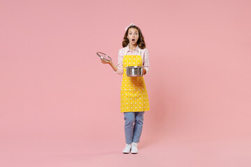 Full length portrait of shocked young brunette woman housewife 20s in yellow apron hold saucepan with lid while doing housework isolated on pastel pink colour background studio. Housekeeping concept.