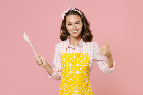 Smiling Young Woman Housewife In Yellow Apron Checkered Shirt Hold Spoon Soup Ladle Dipper Showing Thumb Up Doing Housework Isolated On Pastel Pink Background Studio Portrait. Housekeeping Concept.