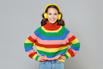 Smiling cheerful funny young brunette woman 20s in casual colorful sweater listening music with headphones standing with arms akimbo looking camera isolated on grey colour background, studio portrait.