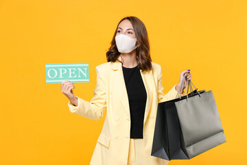 Young woman in suit jacket face mask to safe from coronavirus virus covid-19 hold package bags with purchases after shopping sign with OPEN title isolated on yellow background studio. Black friday.