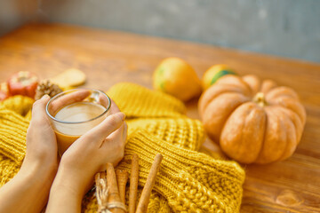 Coffee cup in hand on a knitted sweater. Ripe pumpkins, cinnamon sticks and an orange sweater on a wooden background. Autumn composition for posters and postcards.