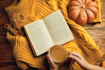 Obraz na płótnie Canvas Hot cocoa in hands, ripe pumpkin, warm sweater and open book on wooden background. Vintage book with romantic stories and fairy tales. Top view of autumn composition with book and knitted sweater.