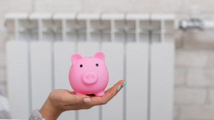 Close up of a female hand holding piggy bank with heating radiator on background