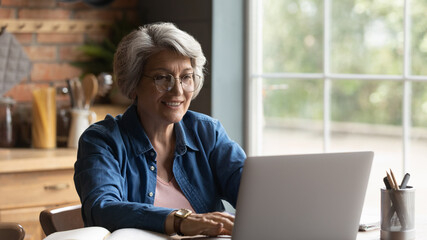 Focused happy elderly 60s grey-haired woman in glasses look at laptop screen work online at home....