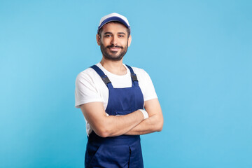 Portrait of happy handyman in overalls and cap standing with crossed hands, profession of service industry, courier delivery. Expert house repairman in workwear smiling. indoor studio shot isolated
