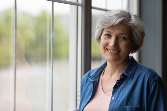 Portrait of smiling mature Caucasian 60s woman stand near window in own house show optimism at retirement. Profile picture of happy elderly female feel positive overjoyed, enjoy good days at home.