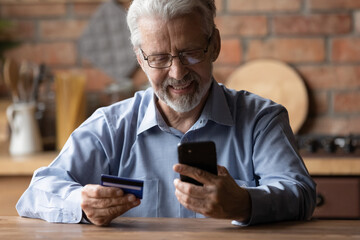 Smiling elderly gray-haired 70s man pay bills online using modern smartphone gadget and credit...