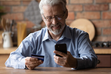 Smiling elderly gray-haired 70s man pay bills online using modern smartphone gadget and credit card. Happy mature bank client make internet payment purchase on cellphone from home. Technology concept.