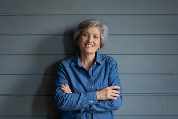 Profile picture of smiling elderly 60s grey-haired woman stand isolated on grey wooden wall...
