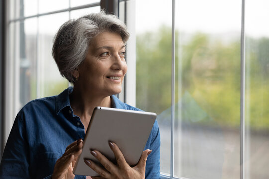 Happy mature grey-haired Caucasian woman use modern tablet gadget look in window distance thinking. Smart senior 60s grandmother browse internet on pad device at home. Elderly and technology concept.