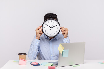 Overtime work. Anonymous man employee sitting in office workplace, hiding face behind clock, sticky notes all around reminding of deadline, stressful job. studio shot isolated on white background