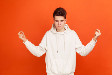 Handsome teenager in casual style sweatshirt holding hands up in mudra gesture closing eyes, practicing yoga, rituals for luck before exams. Indoor studio shot isolated on orange background