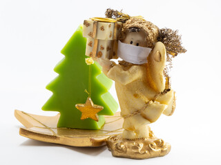 Angel figurine wearing paper mask next to small Christmas tree in front of white background