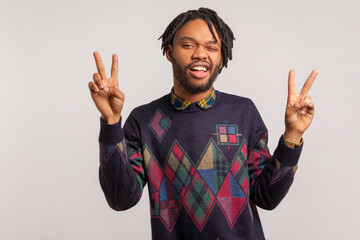 Funny african man with beard and dreadlocks having fun winking and showing peace gesture with fingers with toothy smile on face, teasing. Indoor studio shot isolated on gray background