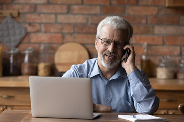 Portrait of smiling mature man sit at table at home talk on cellphone work on computer gadget. Smart senior 70s male have smartphone call conversation, use laptop. Elderly and technology concept.