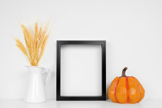 Mock up black frame with fall wheat and pumpkin decor on a white shelf. Autumn concept. Portrait frame against a white wall.