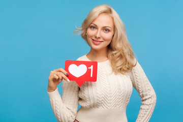 Beautiful blond woman in white knitted sweater with toothy smile on face holding sign with like...