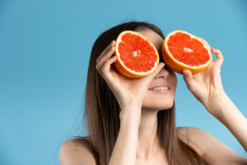 Joyful happy young woman enjoys smell of fresh grapefruit, holding two slices of citrus fruit, cute cheerful girl smiling, posing isolated over blue wall background .