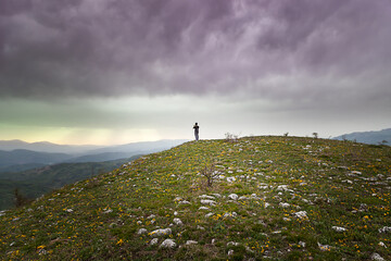 Mountain hiker standing at the edge of the peak coverd by green grass and yellow flowers and photographing distant mountains under a stormy, dark clouds on the sky - obrazy, fototapety, plakaty