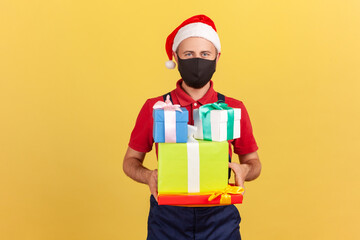 Deliveryman in black protective mask and santa claus hat standing in uniform and holding parcels in hands, delivery service during coronavirus. Indoor studio shot isolated on yellow background