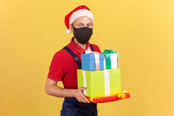 Postman in protective mask uniform and santa claus hat holding and showing parcels and presents, delivery of holidays gifts during quarantine. Indoor studio shot isolated on yellow background