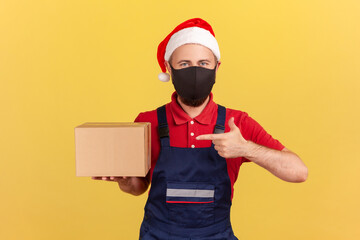 Fototapeta na wymiar Deliveryman in uniform and holiday santa claus hat with protective medical mask pointing finger at cardboard box in his hand, delivery on quarantine. Indoor studio shot isolated on yellow background
