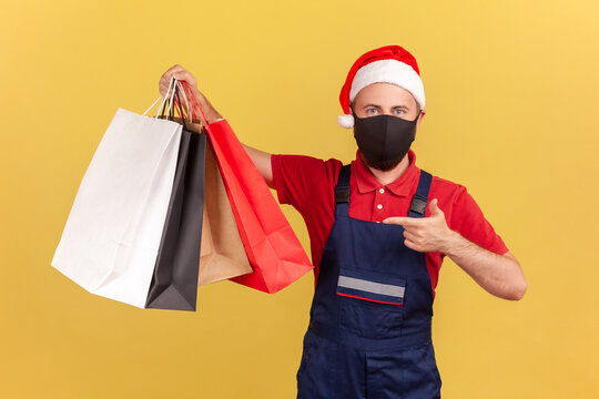 Male Courier In Protective Medical Mask And Holiday Santa Claus Hat Pointing Finger At Shopping Bags In Hand, Easy Delivery During Quarantine. Indoor Studio Shot Isolated On Yellow Background