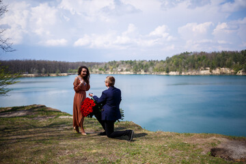 Fototapeta na wymiar Wedding background in the forest: a man made a surprise, a ring gives a beautiful girl a declaration of love and a marriage proposal. Girl with a smile on her face and a bouquet of red roses