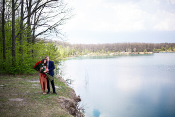 Obraz na płótnie Canvas Cheerful newlyweds go holding hands and laughing, against the background of a lake and a green meadow. Cheerful groom and beautiful bride with curly hair walk in the meadow