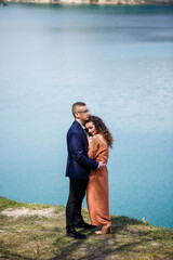 Cheerful newlyweds go holding hands and laughing, against the background of a lake and a green meadow. Cheerful groom and beautiful bride with curly hair walk in the meadow