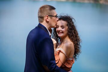 Cheerful newlyweds go holding hands and laughing, against the background of a lake and a green meadow. Cheerful groom and beautiful bride with curly hair walk in the meadow