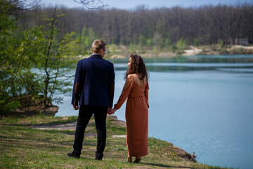 Cheerful newlyweds go holding hands and laughing, against the background of a lake and a green meadow. Cheerful groom and beautiful bride with curly hair walk in the meadow