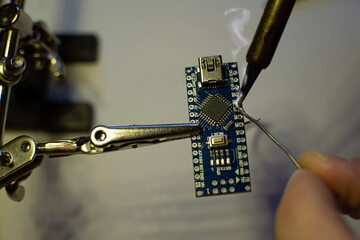 The process of soldering wires to an arduino nano board. The clamp holds the board. There is rosin smoke above the board. Hand holds solder. Soldering iron with a fireproof tip.