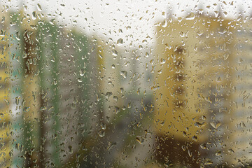 Multicolored buildings behind the misted window glass in rainy day. Blurry background wallpaper with raindrop on windows in raining day. Reflection in drops.
