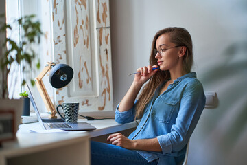 Casual young concentrated woman wearing round glasses working online at a laptop at a comfy...
