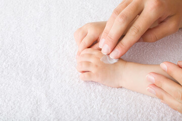 Young mother fingers applying moisturizing cream on baby hand on white towel. Care about children clean and soft body skin. Side view. Closeup.