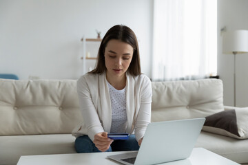 Young Caucasian woman sit on sofa in living room shopping online on laptop with credit card. Millennial female make internet payment on computer, enter bank details, use secure web service system.
