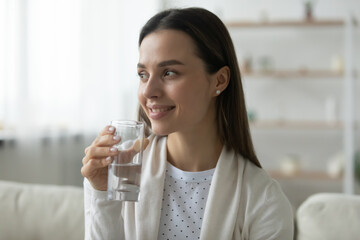 Smiling young Caucasian woman drink pure clean mineral water from glass look in distance. Happy millennial girl feel dehydrated enjoy clear aqua for body refreshment. Hydration, healthy life concept