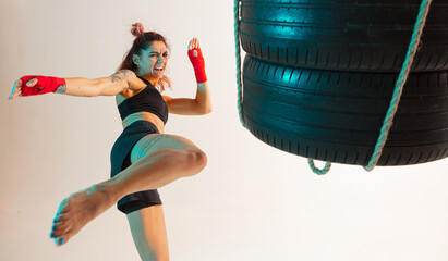 Cool female fighter trains kicking with punching bag made of tires in neon studio light. Women's...