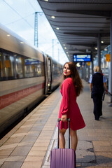 Young attractive woman in rose dress with small violet suitcase on the platform at the railway station going to the train. Travelling inside the country.