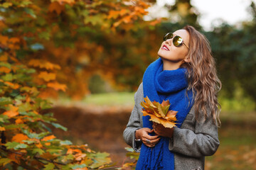 Outdoors lifestyle fashion image of happy pretty girl walking on the sunny autumn park. Holding maple leaves. Wearing stylish grey coat, blue scarf and sunglasses. Smiling and enjoying autumn nature.