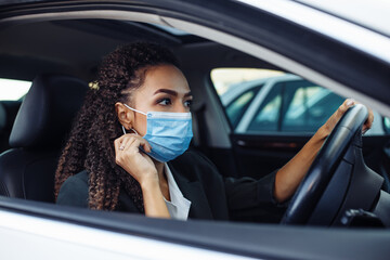 Young business woman fixing and adjusting her medical mask while sitiing in the car behind the steering wheel. Business trips during pandemic, new normal and coronavirus travel safety concept.